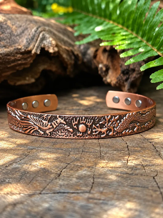 A copper-toned metal cuff bracelet with embossed dragon designs, displayed on a textured wooden surface with natural foliage in the background.