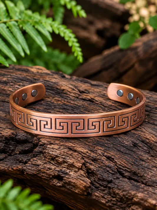 Copper bracelet with a greek key geometric pattern on a natural wooden surface with green ferns in the background 