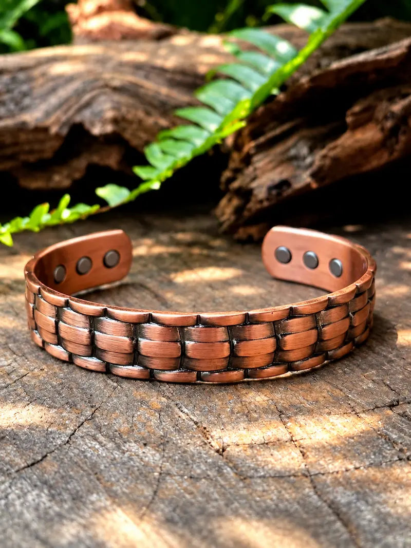 A handmade copper cuff bracelet with a woven link pattern, resting on a rustic wood log with green ferns in the background.
