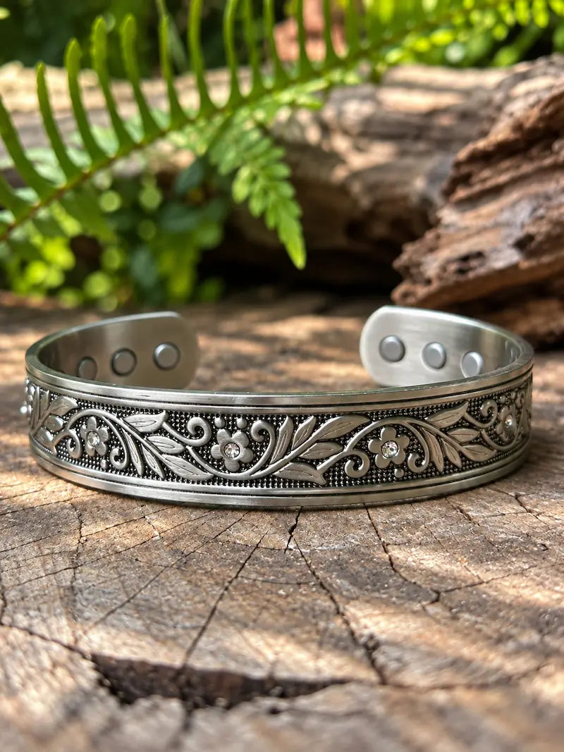 A copper bracelet with a silver finish and a floral design, resting on a natural wooden surface with green ferns in the background 