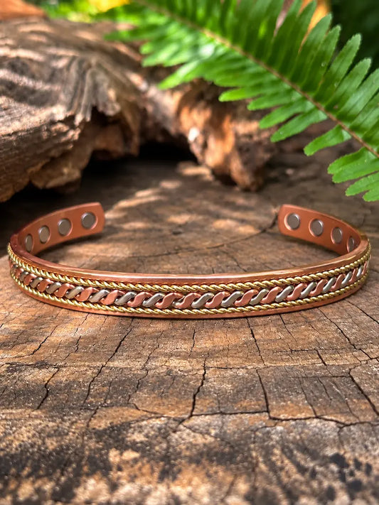 A copper cuff bracelet with a twisted copper and gold metal detailing, displayed on a rustic wood log with a green fern in the background.