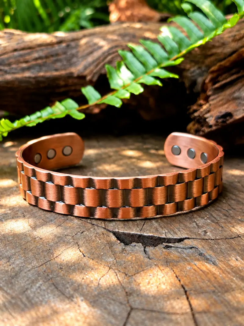 A copper cuff bracelet with a textured woven link pattern, displayed on a rustic wood log with a green fern in the background. 