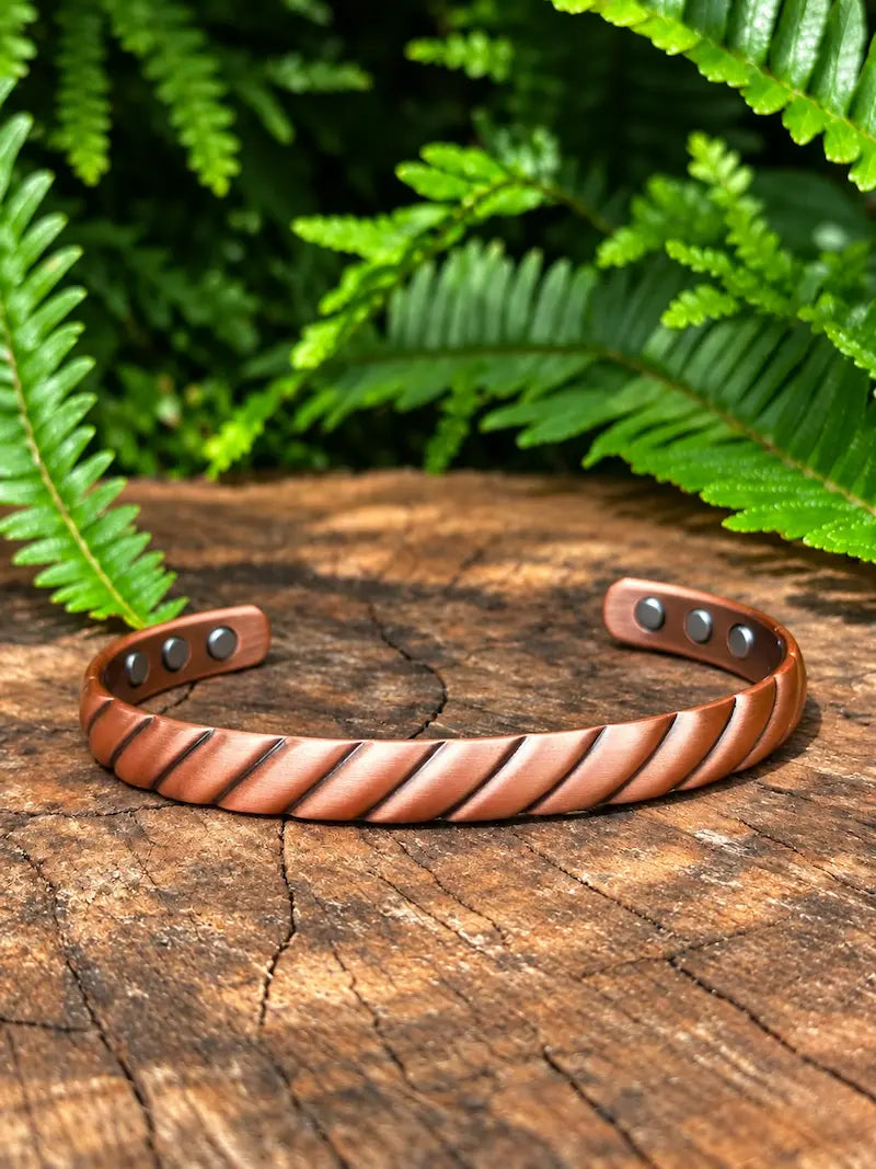 A twisted copper cuff bracelet, displayed on a rustic wooden log with green ferns in the background.