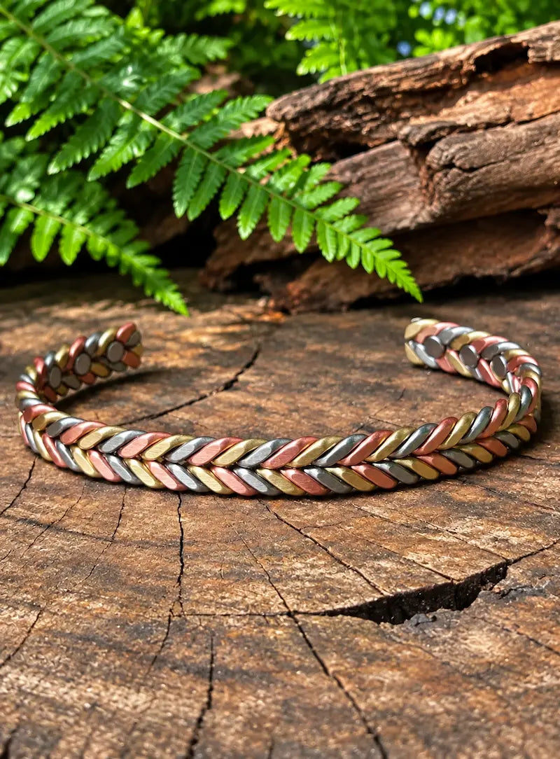 A braided copper cuff bracelet with gold, copper and silver tones, displayed on a rustic tree stump with green ferns in the background.