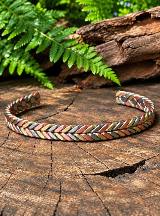 A braided copper cuff bracelet with gold, copper and silver tones, displayed on a rustic tree stump with green ferns in the background.