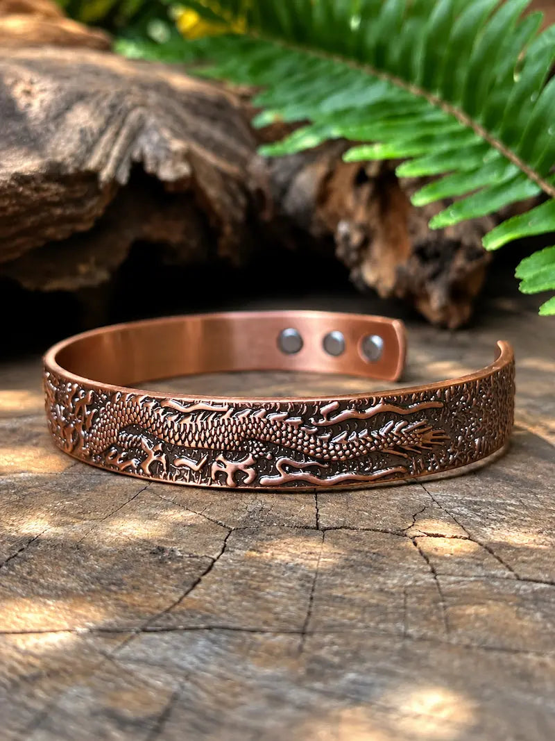 A copper-toned engraved cuff bracelet with a raised dragon motif, displayed on a weathered wood surface with a green fern leaf and dark log in the background.