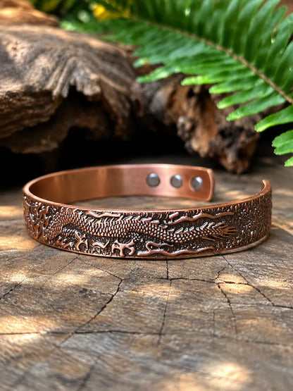 A copper-toned engraved cuff bracelet with a raised dragon motif, displayed on a weathered wood surface with a green fern leaf and dark log in the background.