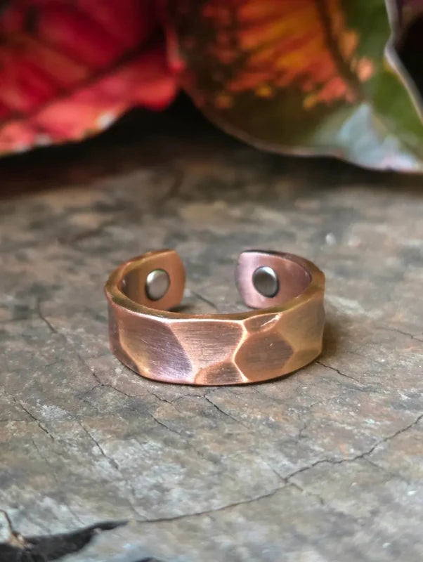 A hammered copper ring resting on a textured gray stone surface with blurred red and green leaves in the background.