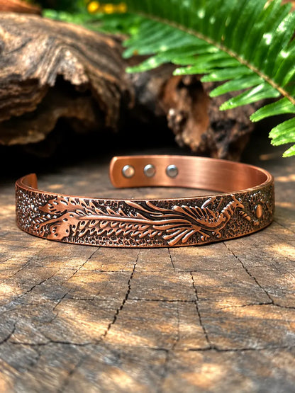 A copper-toned metal cuff bracelet with embossed dragon designs, displayed on a textured wooden surface with natural foliage in the background.