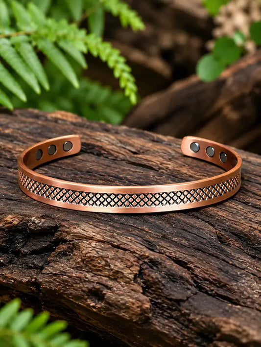 A copper-toned open cuff bracelet with a geometric patterned center band and small magnets at the ends, displayed on a textured log with green foliage in the background.