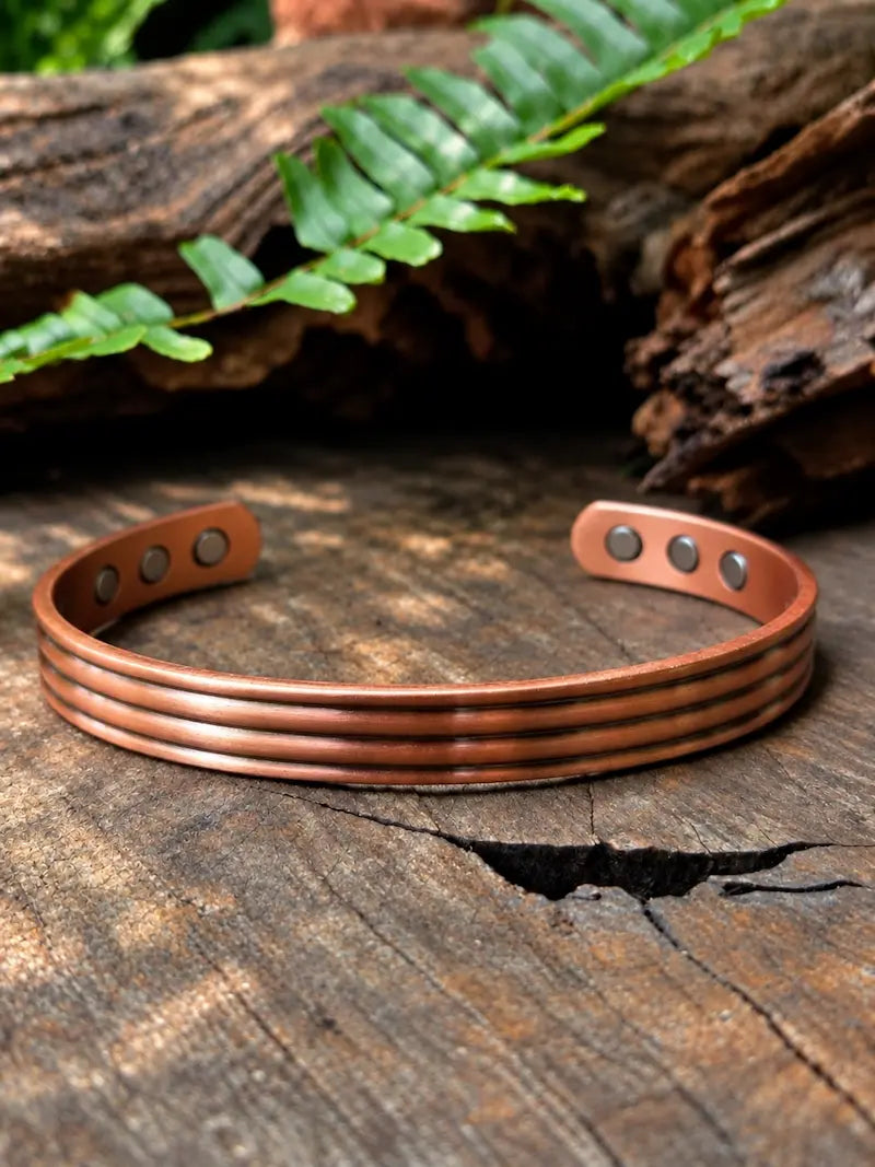 A copper cuff bracelet with a line stripe pattern, displayed on a rustic wooden surface with a green fern leaf in the background.