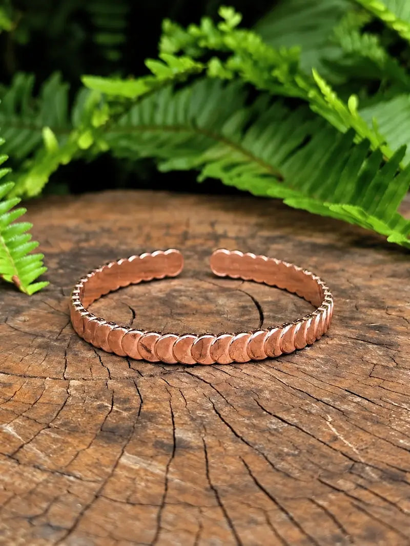 A copper cuff bracelet with a scalloped edge design, displayed on a rustic wood slice with green fern leaves in the background.