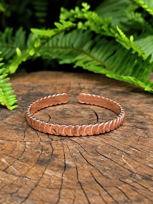 A copper cuff bracelet with a scalloped edge design, displayed on a rustic wood slice with green fern leaves in the background.