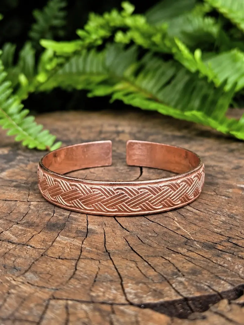 A copper cuff bracelet with an celtic pattern, resting on a rustic wood slice with green ferns in the background.