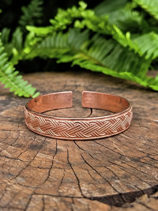 A copper cuff bracelet with an celtic pattern, resting on a rustic wood slice with green ferns in the background.