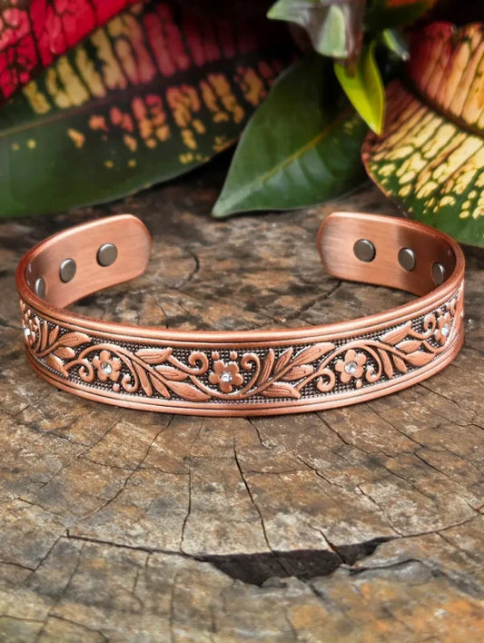 A copper open cuff bracelet with engraved floral scrollwork and small clear rhinestone accents, displayed on a rustic wood surface with blurred green leaves and patterned foliage in the background.
