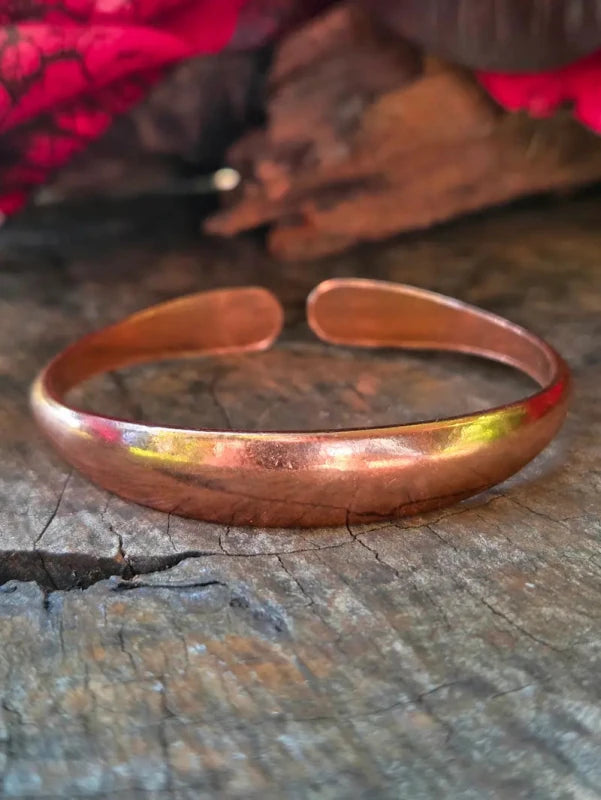 A copper cuff bracelet resting on a rustic wooden surface with a blurred red fabric background.