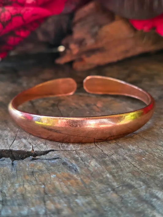 A copper cuff bracelet resting on a rustic wooden surface with a blurred red fabric background.