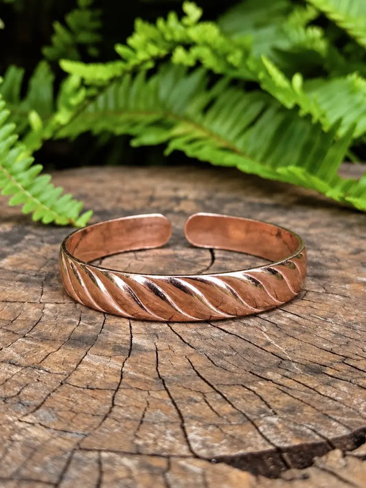 A copper cuff bracelet with a twisted design, resting on a rustic tree stump with green ferns in the background.