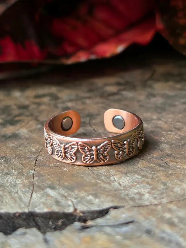 A copper ring with engraved butterfly motifs, photographed on a textured stone surface with a blurred red leaf in the background.