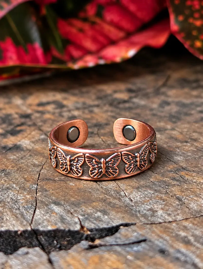 A copper-toned open cuff ring with engraved butterfly motifs, displayed on a textured wooden surface with blurred red leaves in the background.