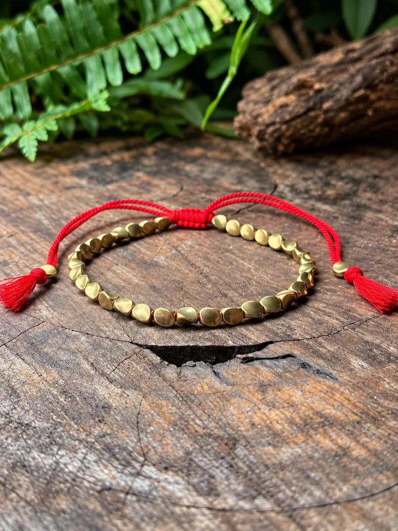 A gold-toned copper beaded bracelet with red cord ties and tassels, displayed on a rustic wooden surface with green fern leaves in the background.